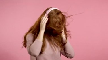 Kızıl saçlı genç kız müzik dinliyor, pembe arka planda stüdyoda kulaklıkla enerjik ve aktif bir şekilde dans ediyor. Müzik, dans, radyo konsepti, yavaş çekim