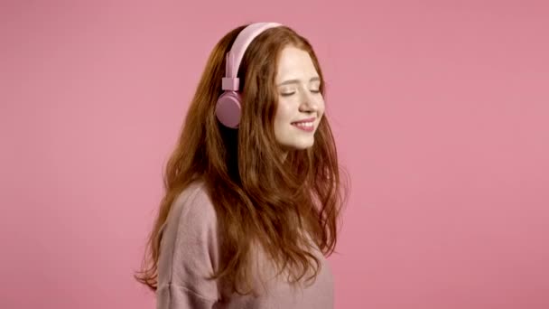 Kızıl saçlı güzel bir kız müzik dinliyor, gülümsüyor, stüdyoda pembe kulaklıklarla dans ediyor. Müzik, dans, radyo konsepti, yavaş çekim. — Stok video