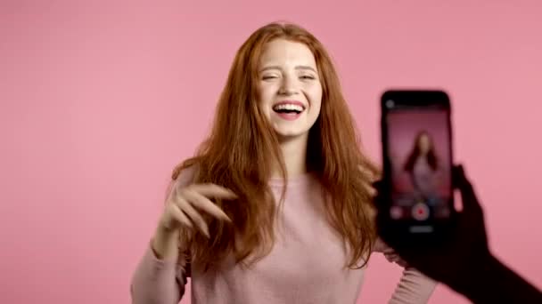 ピンクの背景にスマートフォンのカメラの前で踊っている自分のビデオを記録する笑顔のvloggerの女性。インフルエンサーは面白いソーシャルメディアクリップを作る — ストック動画