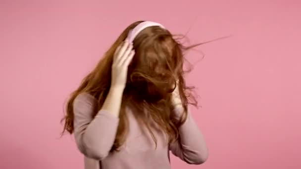 Menina com cabelos vermelhos ouvindo música, dançando energeticamente e ativamente em fones de ouvido em estúdio em fundo rosa. Música, dança, conceito de rádio, câmera lenta — Vídeo de Stock