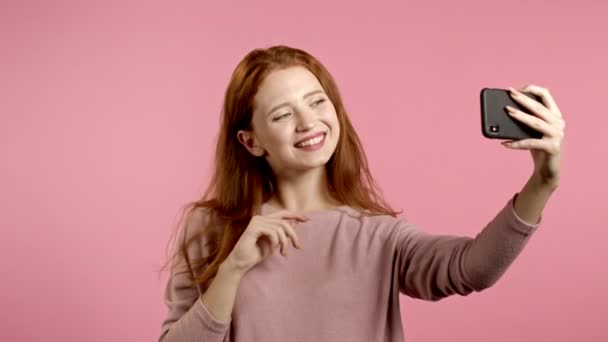 ピンクを背景にスマホで自撮りする笑顔の幸せ女性。テクノロジー、モバイルデバイス、ソーシャルネットワークの概念. — ストック動画
