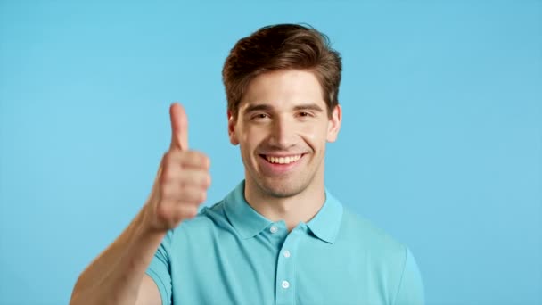 ハンサムな若い男がカメラに微笑む。青い背景の看板のように親指を立てる男。勝者だ。成功だ。ボディランゲージ. — ストック動画