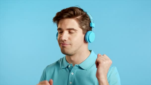 Joven guapo con peinado de moda bailando con auriculares azules en el estudio sobre fondo brillante. Música, danza, concepto de radio, cámara lenta — Vídeo de stock