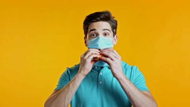 Молодий чоловік у захисній медичній масці їсть гамбургер на жовтому студійному тлі. Хлопець розважається, показує, як жити під час епідемії. — стокове відео