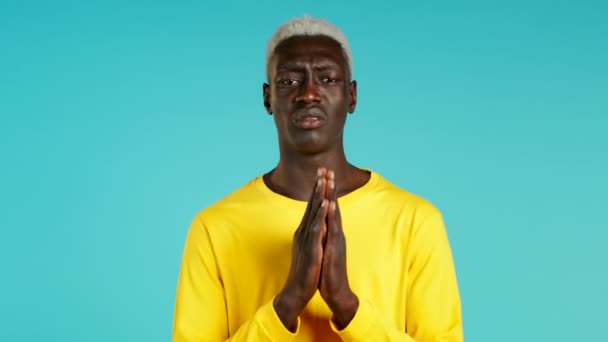 Mladý africký Američan ve žlutém tričku se modlí nad modrým pozadím. Chlápek, který někoho prosí, aby uspokojil jeho touhy.. — Stock video