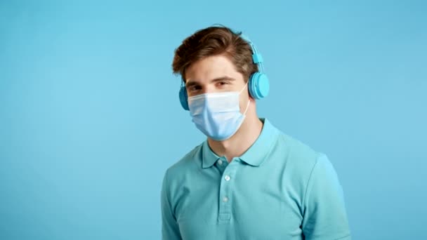 Beau jeune homme avec masque de protection s'amusant, souriant, tête de danse avec écouteurs bleus en studio. Musique, danse, concept radio. — Video