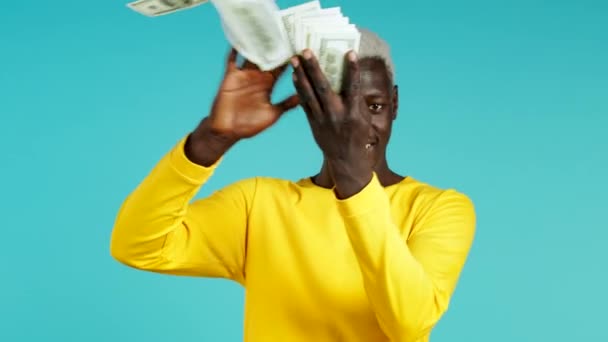 African man with surprised happy face scatters, waste money. Person overspend US currency. Guy in yellow is flush with dollars on blue studio background . — Stock Video