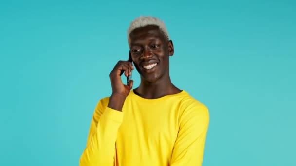 Νεαρός μαύρος Αφρικανός μιλάει στο κινητό του σε μπλε φόντο. Ο μοδάτος τύπος με τα κίτρινα έχει συζήτηση. Smartphone, έννοια της τεχνολογίας. — Αρχείο Βίντεο