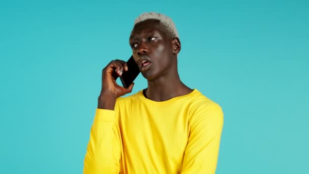 Joven africano negro hablando por teléfono móvil sobre fondo azul. Chico de moda en amarillo tienen conversación. Smartphone, concepto de tecnología. — Vídeos de Stock