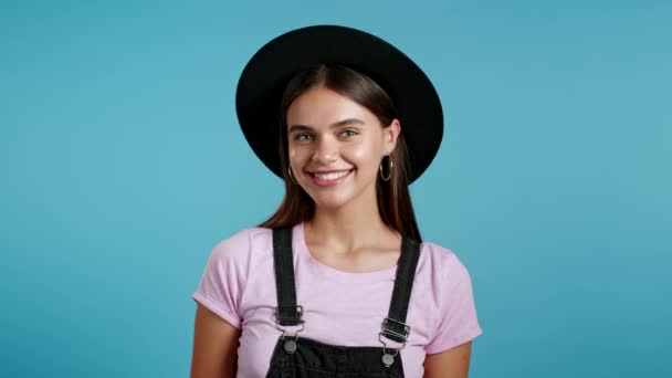 Jonge lachende vrouw met hoed die naar de camera kijkt. Portret van schattig mooi hipster meisje op blauwe studio muur achtergrond. — Stockvideo