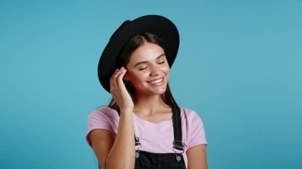 Atractiva mujer hipster juguetona en el desgaste general y el sombrero sonriendo, coqueteando con la cámara sobre fondo azul de la pared. Lindo retrato de chicas de moda — Vídeo de stock