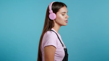 Güzel genç kız müzik dinliyor, gülümsüyor, pembe kulaklıklarla dans ediyor. Stüdyoda mavi arka planda. Müzik, dans, radyo konsepti, yavaş çekim.