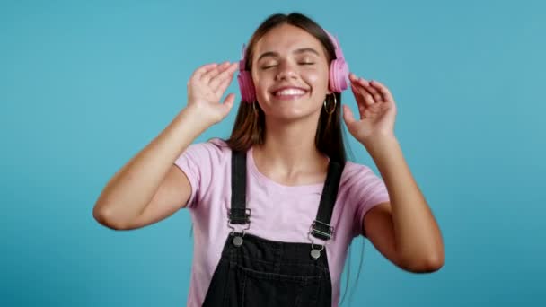 Chica joven y bonita con el pelo largo escuchando música, sonriendo, bailando las manos en auriculares rosados en el estudio sobre fondo azul. Música, danza, concepto de radio, cámara lenta. — Vídeos de Stock