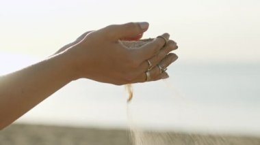 Kumsalda parmaklarının arasından boho halkalarıyla kum döken bir kadının güneş ışığıyla yavaş çekim halkalarına yakın çekim. Güneşin doğuşunda ya da batışında kumla oynayan güzel bir bayan sahnesi.