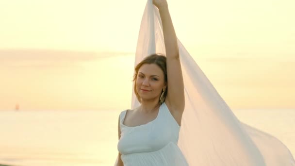 Красивая женщина стоит на морском пляже и держит длинную белую ткань шаль, как паруса, развевающиеся на ветру. Понятие женственности, нежности, свободы. — стоковое видео