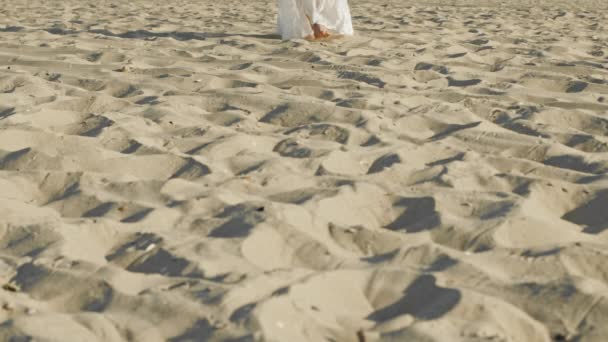Piernas de niña vestida de blanco vestido largo caminando descalzo arena en la playa del mar o el desierto. En cámara lenta. Mujer camina a cámara — Vídeo de stock