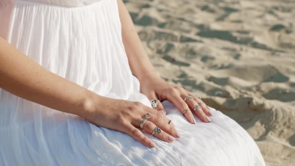 Donna irriconoscibile in abito bianco e anelli boho sulle braccia siede solo sulla spiaggia di sabbia. Meditazione, immersione nel mondo interiore, unità con la natura. — Video Stock
