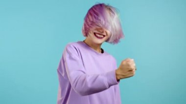 Mor boyalı saçı olan alışılmadık bir kadın çok mutlu ve mutlu, zafere evet işareti yapıyor, sonuca ulaştı, amaçlarına ulaştı. Mavi arka planda heyecanlı mutlu bir kız.