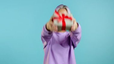 Renkli saçlı, elinde fiyonklu hediye kutusu olan ve mavi arka plandaki kameraya elleriyle veren hoş bir genç kadın. Kız gülümsüyor, o mutlu..
