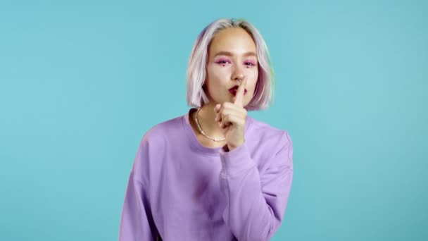 Dziewczyna z fioletową fryzurą trzymając palec na ustach na niebieskim tle. Cisza, tajemnica, cisza. Zamknij się.. — Wideo stockowe