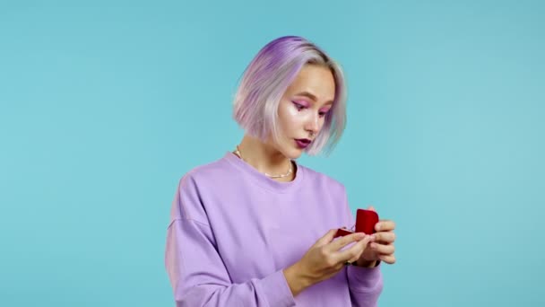 Jovem mulher atraente com cabelo colorido tingido segurando pequena caixa de jóias com anel de diamante proposta no fundo da parede azul. Senhora sorrindo, ela está feliz por estar presente, proposta de casamento — Vídeo de Stock