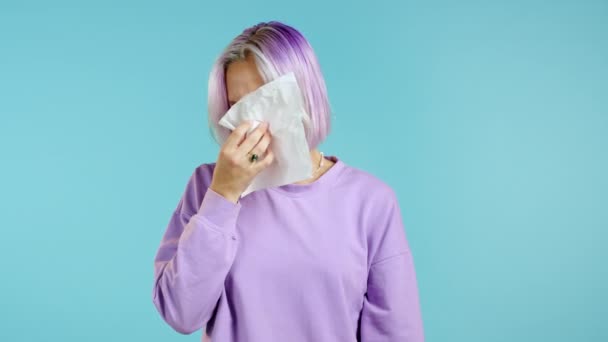 Необычная женщина с фиолетовыми волосами чихает в ткань. Изолирован на синем фоне студии. Девушка больна, у нее простуда или аллергическая реакция. Коронавирус, эпидемия 2020 года, концепция болезни — стоковое видео