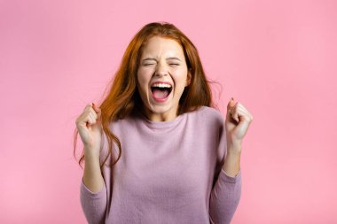 Kızıl saçlı kadın çok mutlu ve mutlu. Zaferin evet jestini gösteriyor. Sonuç elde etti. Pembe arka planda heyecanlı ve mutlu bir bayan.