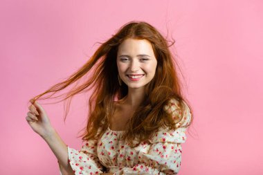 Uzun kırmızı dalgalı, parlak saçlı, kameraya bakıp flört eden genç bir kadın. Güzel model kız zarif gür saçlarını gösteriyor. Pembe stüdyo arka planı.