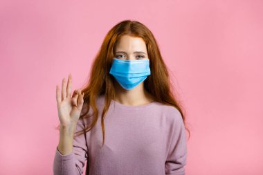 Pembe arka planda tıbbi maskeli mutlu kadın. Uzun kızıl saçlı bayan iyi bir jest gösteriyor. Kazandım. Başarı. Vücut dili.