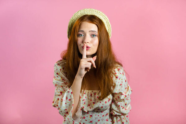 Smiling woman with red hair holding finger on her lips over pink background. Gesture of shhh, secret, silence. Close up.