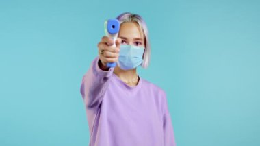 Koruyucu maskeli kadın, elektronik temas olmayan termometreyi kamera merceğine yönlendirir ve sıcaklığı ölçer. Jestle her şeyin yolunda olduğunu, sıcaklığın normal olduğunu gösterir. Mavi stüdyo arka planı
