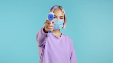 Koruyucu maskeli kadın, elektronik temas olmayan termometreyi kamera merceğine yönlendirir ve sıcaklığı ölçer. Sıcaklığın yüksek olduğunu jestle gösteriyor. Mavi stüdyo arka planı