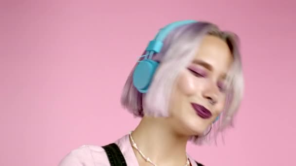 Retrato de cerca de una chica bonita con el pelo teñido de violeta escuchando música, sonriendo, bailando en auriculares en el estudio sobre fondo rosa. Música, danza, concepto de radio, cámara lenta. — Vídeo de stock