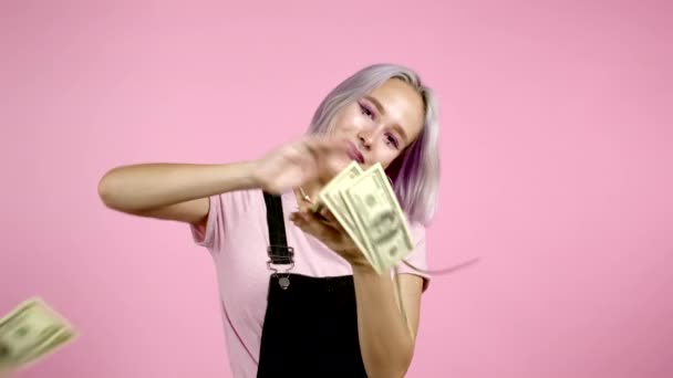 Jolie femme au visage heureux se disperse, gaspille de l'argent. Hipster fille avec les cheveux teints violets surdépenser la monnaie américaine. Dame est affleurant avec des dollars sur fond studio rose . — Video