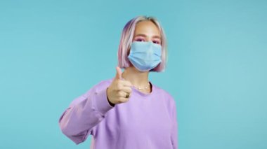 Tıbbi maskesi ve mor boyalı saç stili olan hoş bir kadın mavi arka planda baş parmak işareti gösteriyor. Olumlu genç kız kameraya gülümsüyor. Kazandım. Başarı. Vücut dili. 