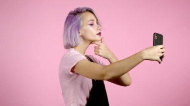 Modayı takip eden kız pembe arka planda akıllı telefondan selfie çekiyor. Menekşe rengi saç stili olan kadın. Teknoloji, mobil cihaz, sosyal ağlar kavramı