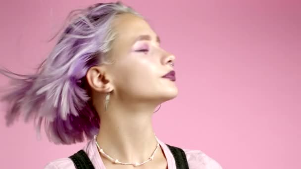 Atractiva mujer hipster con el pelo púrpura colorido y maquillaje brillo sonriente, sacudiendo la cabeza cerca de soplador de viento sobre fondo rosa. Linda chica de moda con peinado ventoso — Vídeo de stock