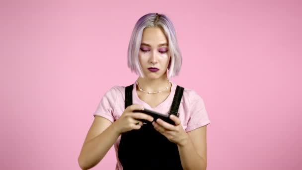 外表不寻常的女孩在粉色工作室墙上的智能手机上玩游戏。使用现代技术-应用、社交网络 — 图库视频影像