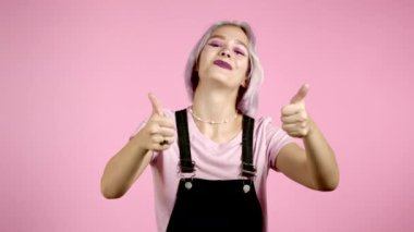 Menekşe boyalı saçlı hoş bir kadın pembe arka planda baş parmak işareti gösteriyor. Olumlu genç kız kameraya gülümsüyor. Kazandım. Başarı. Vücut dili. 