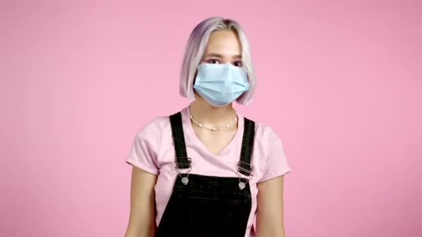 Jonge onzekere vrouw met een beschermend masker haalt armen op, maakt gebaar van ik weet het niet, kan niets helpen. Roze studio achtergrond. — Stockvideo