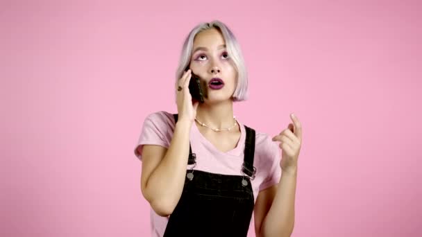 Mujer joven y bonita con el pelo violeta habla en serio por teléfono. Hermosa chica sosteniendo y utilizando el teléfono inteligente. Fondo de estudio rosa. — Vídeos de Stock