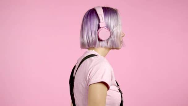 Vista lateral. Chica bonita con el pelo teñido violeta escuchando música, sonriendo, bailando en auriculares en estudio sobre fondo rosa. Música, danza, concepto de radio, cámara lenta. — Vídeo de stock