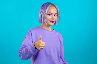 Mor boyalı saç stili olan hoş bir kadın mavi arka planda baş parmak işareti gösteriyor. Olumlu genç kız kameraya gülümsüyor. Kazandım. Başarı. Vücut dili. 