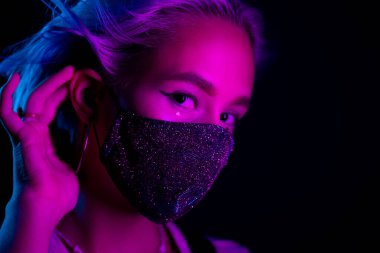 Gece kulübünde sahte elmaslarla dans eden koruyucu maskeli, güzel, modaya uygun genç bir kadın. Neon renkli ışık. Moda modelinin yakın plan portresi.