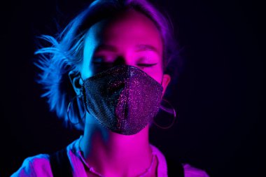 Gece kulübünde sahte elmaslarla dans eden koruyucu maskeli, güzel, modaya uygun genç bir kadın. Neon renkli ışık. Moda modelinin yakın plan portresi.
