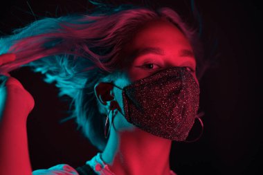 Gece kulübünde sahte elmaslarla dans eden koruyucu maskeli, güzel, modaya uygun genç bir kadın. Neon renkli ışık. Moda modelinin yakın plan portresi.