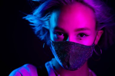 Gece kulübünde sahte elmaslarla dans eden koruyucu maskeli, güzel, modaya uygun genç bir kadın. Neon renkli ışık. Moda modelinin yakın plan portresi.
