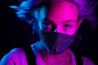Gece kulübünde sahte elmaslarla dans eden koruyucu maskeli, güzel, modaya uygun genç bir kadın. Neon renkli ışık. Moda modelinin yakın plan portresi.