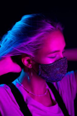 Gece kulübünde sahte elmaslarla dans eden koruyucu maskeli, güzel, modaya uygun genç bir kadın. Neon renkli ışık. Moda modelinin yakın plan portresi.