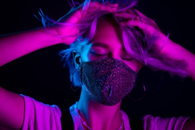 Gece kulübünde sahte elmaslarla dans eden koruyucu maskeli, güzel, modaya uygun genç bir kadın. Neon renkli ışık. Moda modelinin yakın plan portresi.